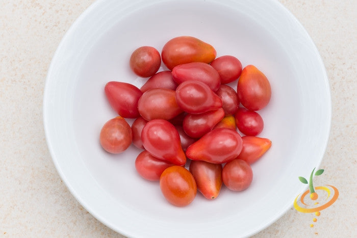 Red pear-shaped indeterminate tomato seeds from SeedsNow, shown in close-up with vibrant green foliage in the background.